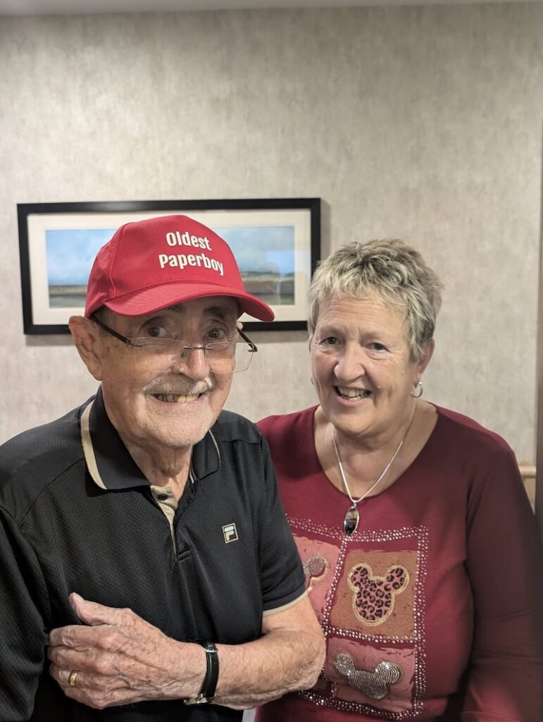 male resident wearing a black polo top smiling wearing a 'oldest paperboy' cap
