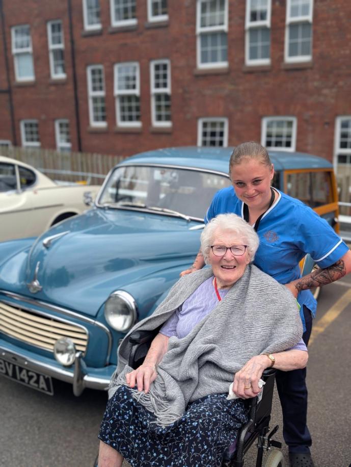 older lady in a wheelchair and a carer in front of a blue car