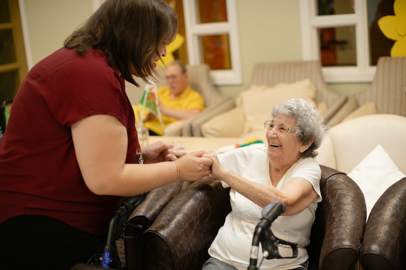 a resident and carer holding hands and smiling in a care home lounge